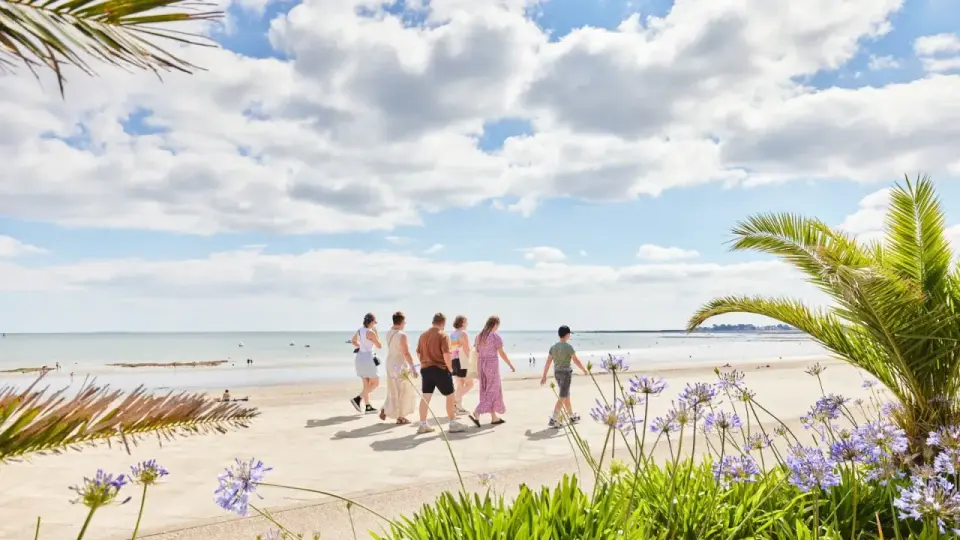 Famille profitant des vacances scolaires en Bretagne, marchant sur la promenade face à la mer, avec des palmiers et des fleurs bleues.